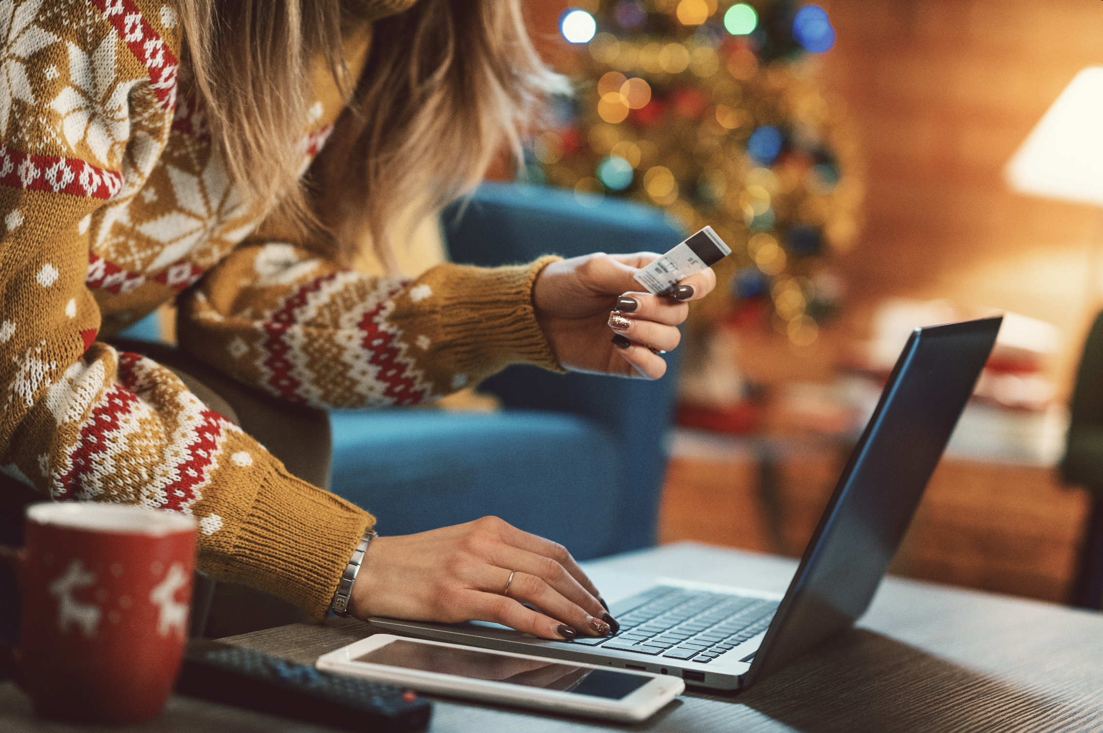 Woman using her credit card to make a online purchase wearing a holiday sweater in front of a Christmas tree