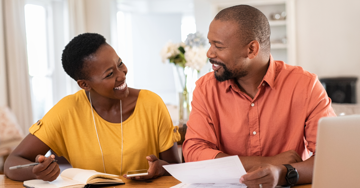 A man and woman smiling while using computer and writing on paper