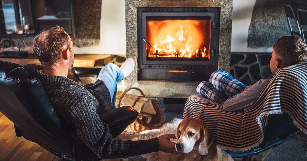 Two men and a dog sitting by a fire in a fireplace